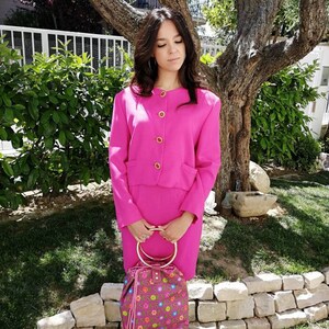 Può includere: Una donna indossa un tailleur gonna rosa acceso con bottoni dorati e una borsa con motivo floreale e manico ad anello dorato.