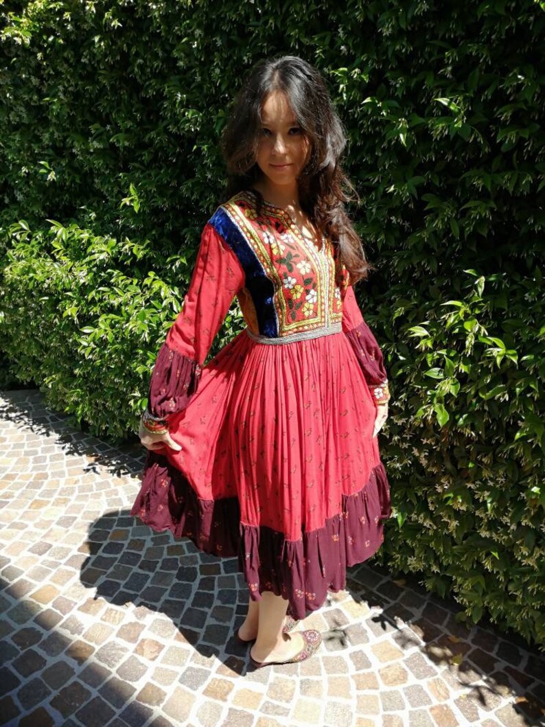 Puede incluir: Una mujer con un vestido rojo y burdeos con bordados intrincados y un ribete de terciopelo azul. El vestido tiene una falda fluida y mangas largas. Est&aacute; de pie frente a un arbusto verde.