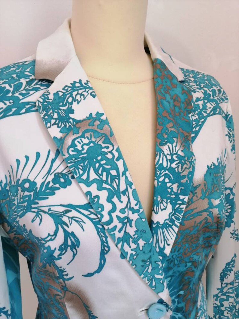May include: A white blazer with a teal and silver floral pattern. The blazer has a single button closure and a notched collar.