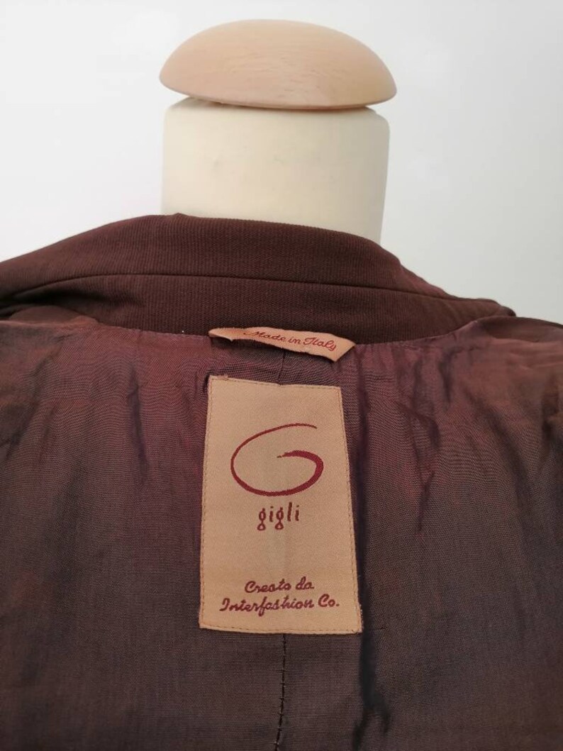 Pu&ograve; includere: Giacca marrone con un'etichetta in pelle marrone che dice "Made in Italy" e "Gigli" con un logo "G" stilizzato. L'etichetta dice anche "Creato da Interfashion Co."