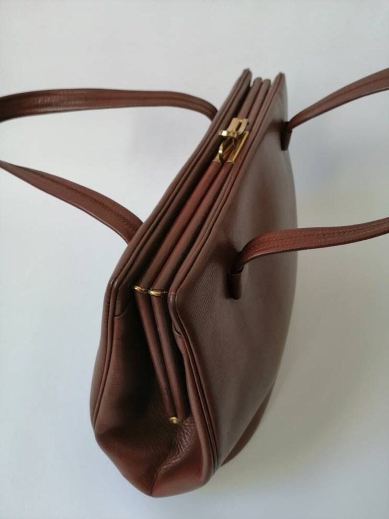 Puede incluir: Un bolso de mano de cuero marr&oacute;n con un cierre de broche dorado y dos asas. El bolso est&aacute; cerrado y tiene una forma ligeramente redondeada.