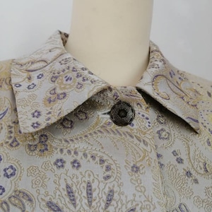 May include: A close-up of a light gray jacket with a paisley pattern in gold and purple. The jacket has a collar and a single button closure.