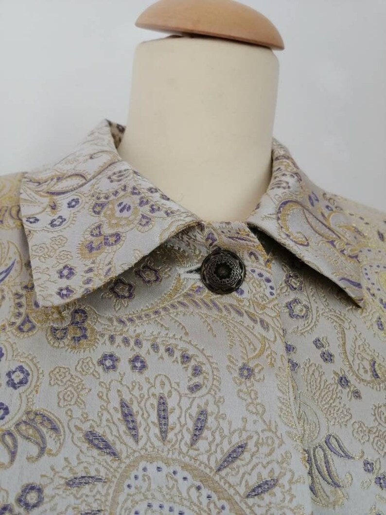 May include: A close-up of a light gray jacket with a paisley pattern in gold and purple. The jacket has a collar and a single button closure.