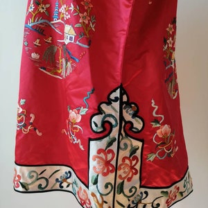 May include: A red silk dress with intricate embroidery featuring a pagoda, flowers, and a small house. The dress has a white trim with black edging and a floral design.