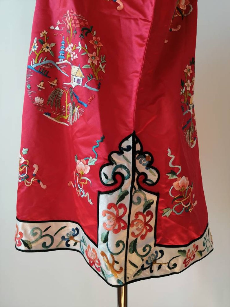 May include: A red silk dress with intricate embroidery featuring a pagoda, flowers, and a small house. The dress has a white trim with black edging and a floral design.