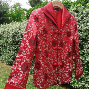 Pu&ograve; includere: Una giacca di seta rossa con un design cinese tradizionale. La giacca ha un colletto alla coreana, una chiusura con bottoni e maniche lunghe. Il tessuto &egrave; decorato con un motivo floreale e dettagli dorati.