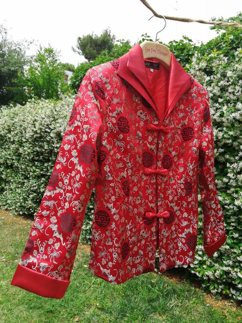 Pu&ograve; includere: Una giacca di seta rossa con un design cinese tradizionale. La giacca ha un colletto alla coreana, una chiusura con bottoni e maniche lunghe. Il tessuto &egrave; decorato con un motivo floreale e dettagli dorati.