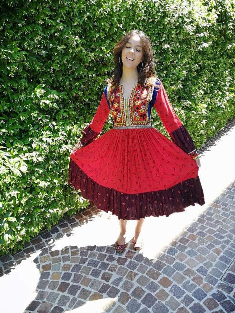 Puede incluir: Una mujer con un vestido rojo y marr&oacute;n con bordados y ribete de terciopelo azul. El vestido tiene una falda larga con un dobladillo con volantes. Est&aacute; de pie en un camino de piedra frente a un seto verde.