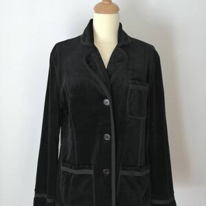 Può includere: Un blazer in velluto nero con chiusura a bottoni e due tasche a toppa. Il blazer ha un bordo nero a contrasto intorno al colletto, alle tasche e ai polsini.