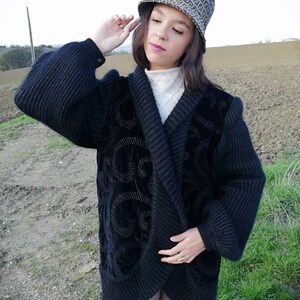 May include: A black knitted cardigan with a velvet design. The cardigan has a shawl collar and long sleeves. The model is wearing the cardigan with a white turtleneck and brown corduroy pants. The model is also wearing a brown and black bucket hat with a pattern.