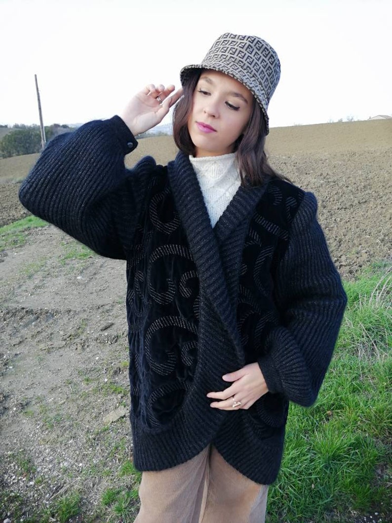 May include: A black knitted cardigan with a velvet design. The cardigan has a shawl collar and long sleeves. The model is wearing the cardigan with a white turtleneck and brown corduroy pants. The model is also wearing a brown and black bucket hat with a pattern.