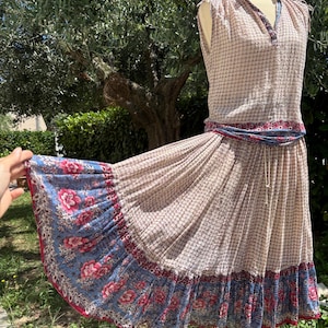 Könnte beinhalten: Ein ärmelloses Kleid im Vintage-Stil auf einer Schaufensterpuppe. Das Kleid hat ein cremefarbenes Oberteil mit kleinem Gittermuster und einen blauen und roten Blumenrock. Ein blauer Schärpe betont die Taille. Das Kleid wird im Freien gezeigt.