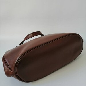 Puede incluir: Un bolso tote de cuero marr&oacute;n con un solo asa. El bolso est&aacute; tumbado de espaldas con la parte inferior del bolso mirando hacia la c&aacute;mara.