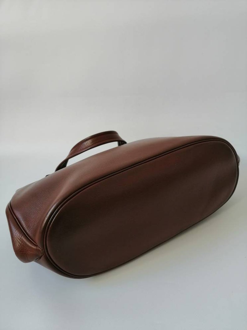 Puede incluir: Un bolso tote de cuero marr&oacute;n con un solo asa. El bolso est&aacute; tumbado de espaldas con la parte inferior del bolso mirando hacia la c&aacute;mara.