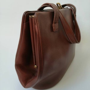 Puede incluir: Un bolso de mano de cuero marr&oacute;n con cierre de solapa y dos asas. El bolso tiene una forma ligeramente redondeada y un acabado de cuero texturizado.