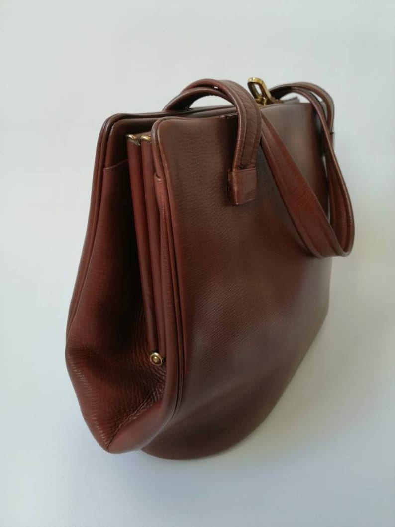 Puede incluir: Un bolso de mano de cuero marr&oacute;n con cierre de solapa y dos asas. El bolso tiene una forma ligeramente redondeada y un acabado de cuero texturizado.