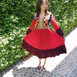 Puede incluir: Un vestido rojo y negro con bordados florales y una falda larga. El vestido tiene cuello en V y mangas largas. La modelo lleva el vestido en un entorno de jard&iacute;n.