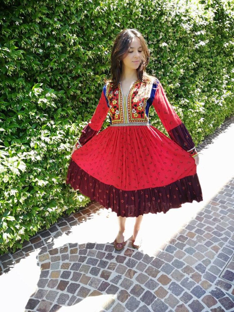 Puede incluir: Un vestido rojo y negro con bordados florales y una falda larga. El vestido tiene cuello en V y mangas largas. La modelo lleva el vestido en un entorno de jard&iacute;n.