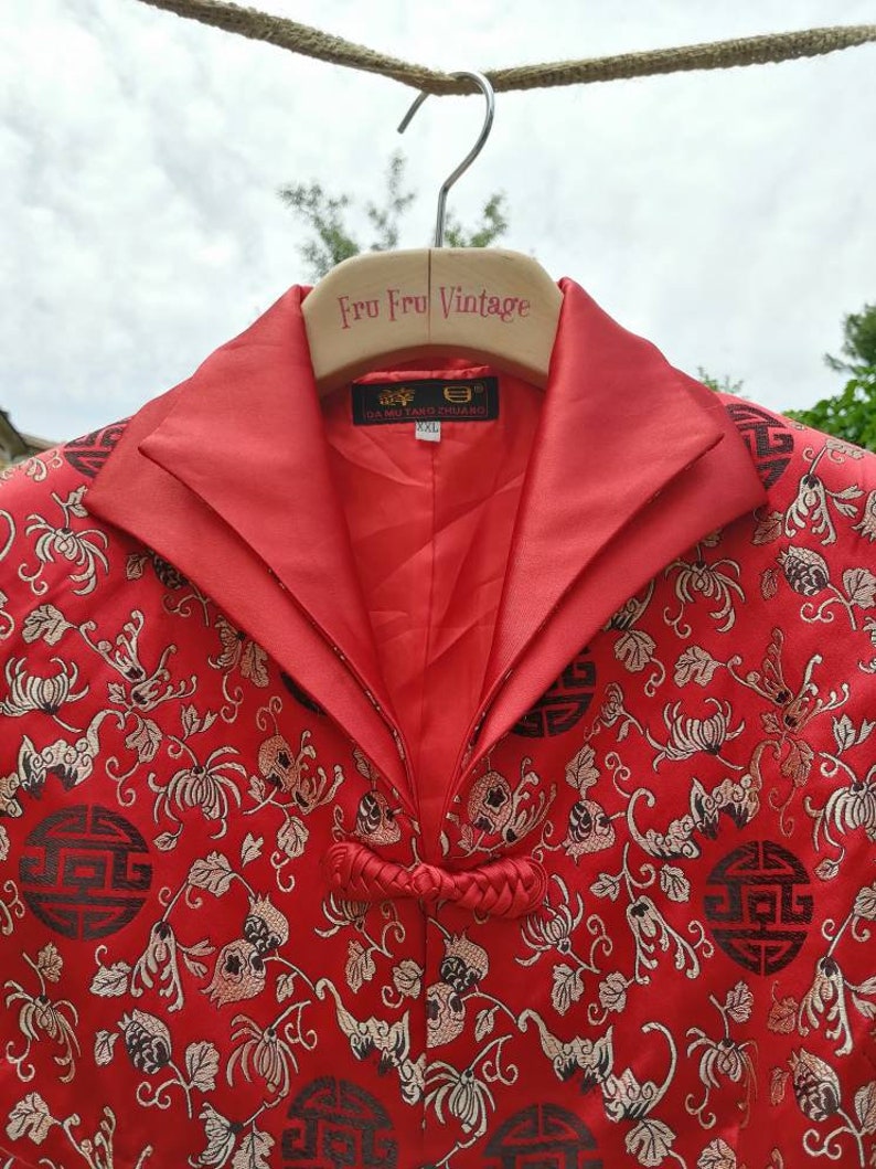 Pu&ograve; includere: Giacca di seta rossa con motivo floreale e design di caratteri cinesi in nero e oro. La giacca ha un colletto alla coreana e una chiusura annodata. L'etichetta sulla giacca recita "DA MU TANG ZHU LIANG" e "XXL".