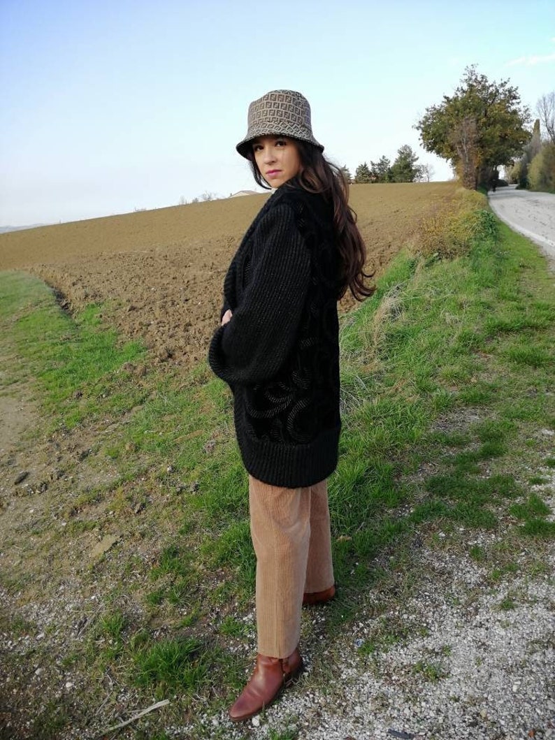 May include: A woman wearing a black sweater, brown corduroy pants, and brown boots. She is wearing a brown and black patterned bucket hat. She is standing in a field with a dirt road in the background.