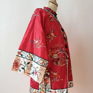 May include: A red silk jacket with intricate floral embroidery and a white trim. The jacket has a mandarin collar and a front closure with a button. The embroidery features flowers, birds, and a pagoda.