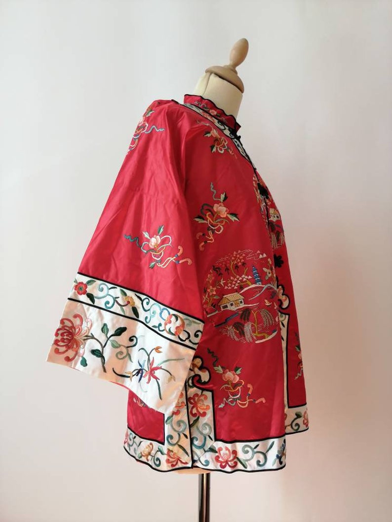 May include: A red silk jacket with intricate floral embroidery and a white trim. The jacket has a mandarin collar and a front closure with a button. The embroidery features flowers, birds, and a pagoda.