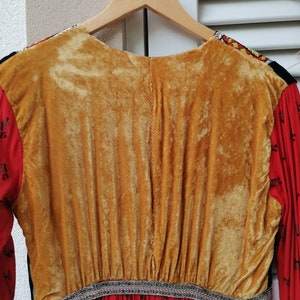 Puede incluir: Un vestido de manga larga con tejido estampado en rojo, negro y blanco. El vestido tiene un panel de terciopelo dorado en la espalda y un ribete negro y plateado alrededor de la cintura.