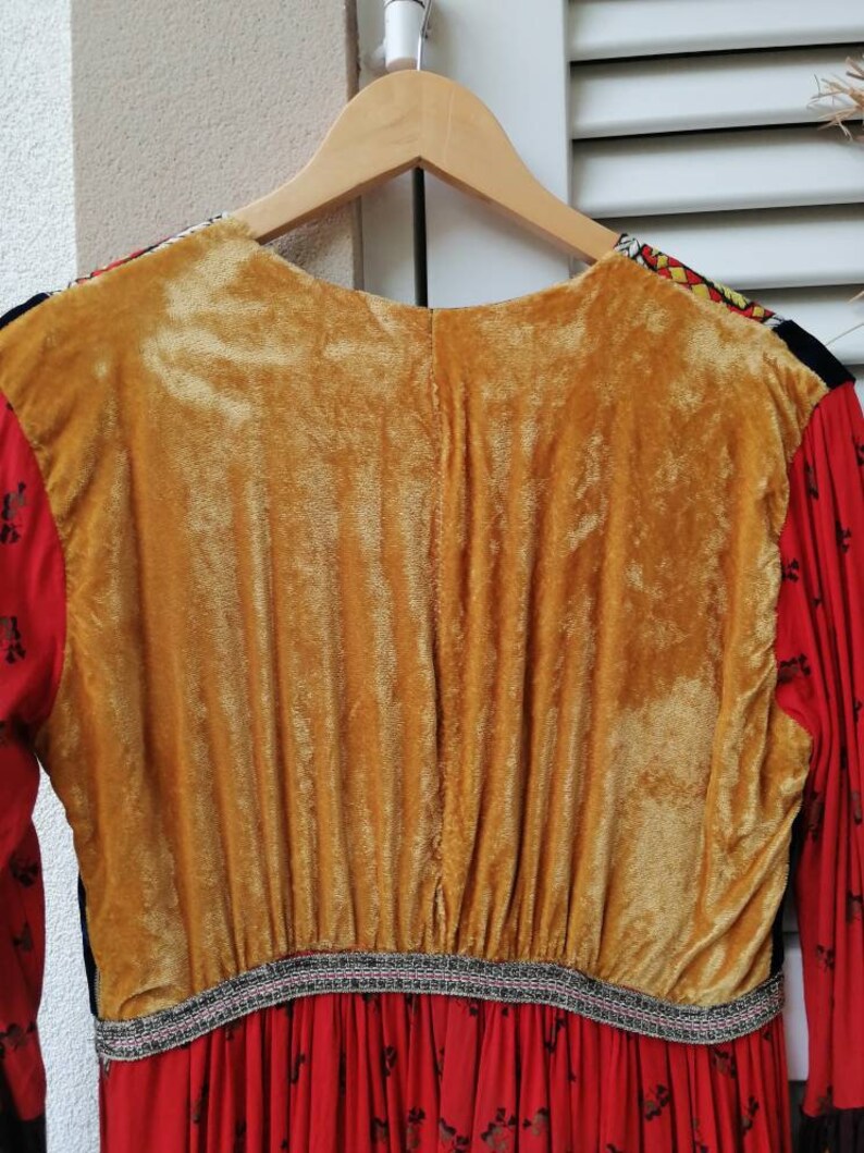 Puede incluir: Un vestido de manga larga con tejido estampado en rojo, negro y blanco. El vestido tiene un panel de terciopelo dorado en la espalda y un ribete negro y plateado alrededor de la cintura.