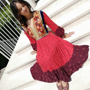 Puede incluir: Una mujer con un vestido rojo y burdeos con bordados florales y ribete de terciopelo azul. Est&aacute; sentada en un conjunto de escaleras blancas.