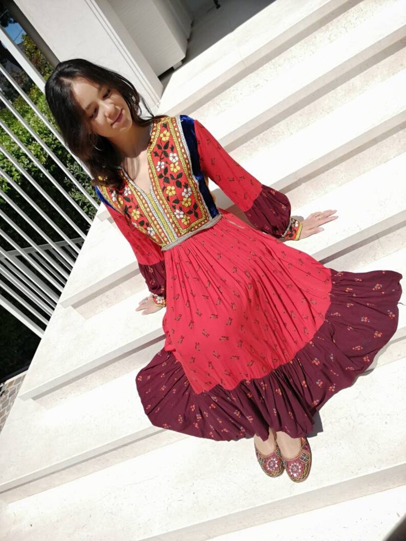 Puede incluir: Una mujer con un vestido rojo y burdeos con bordados florales y ribete de terciopelo azul. Est&aacute; sentada en un conjunto de escaleras blancas.
