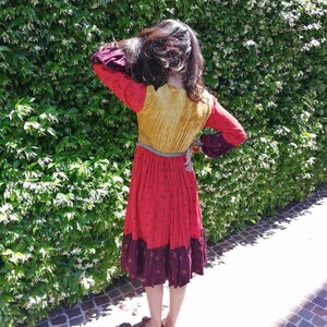 Puede incluir: Una mujer lleva un vestido largo multicolor con un corpi&ntilde;o amarillo, una falda roja y un ribete burdeos. El vestido tiene un dobladillo con volantes y mangas largas. Est&aacute; de pie frente a un seto verde.