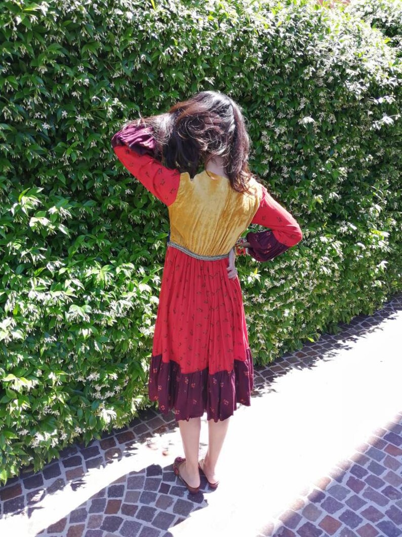Puede incluir: Una mujer lleva un vestido largo multicolor con un corpi&ntilde;o amarillo, una falda roja y un ribete burdeos. El vestido tiene un dobladillo con volantes y mangas largas. Est&aacute; de pie frente a un seto verde.