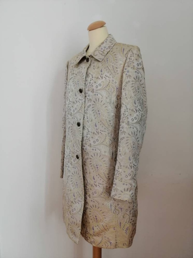 May include: A long-sleeved, beige coat with a paisley pattern. The coat has a button-up front and a collared neckline.