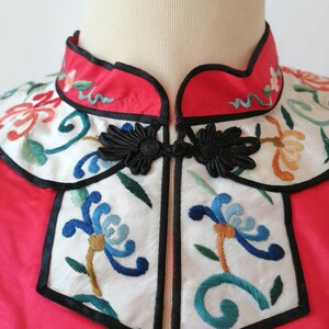 May include: A red Chinese-style jacket with a white embroidered collar and front panel. The collar and front panel feature floral designs in blue, green, orange, and white. The jacket has black frog closures.