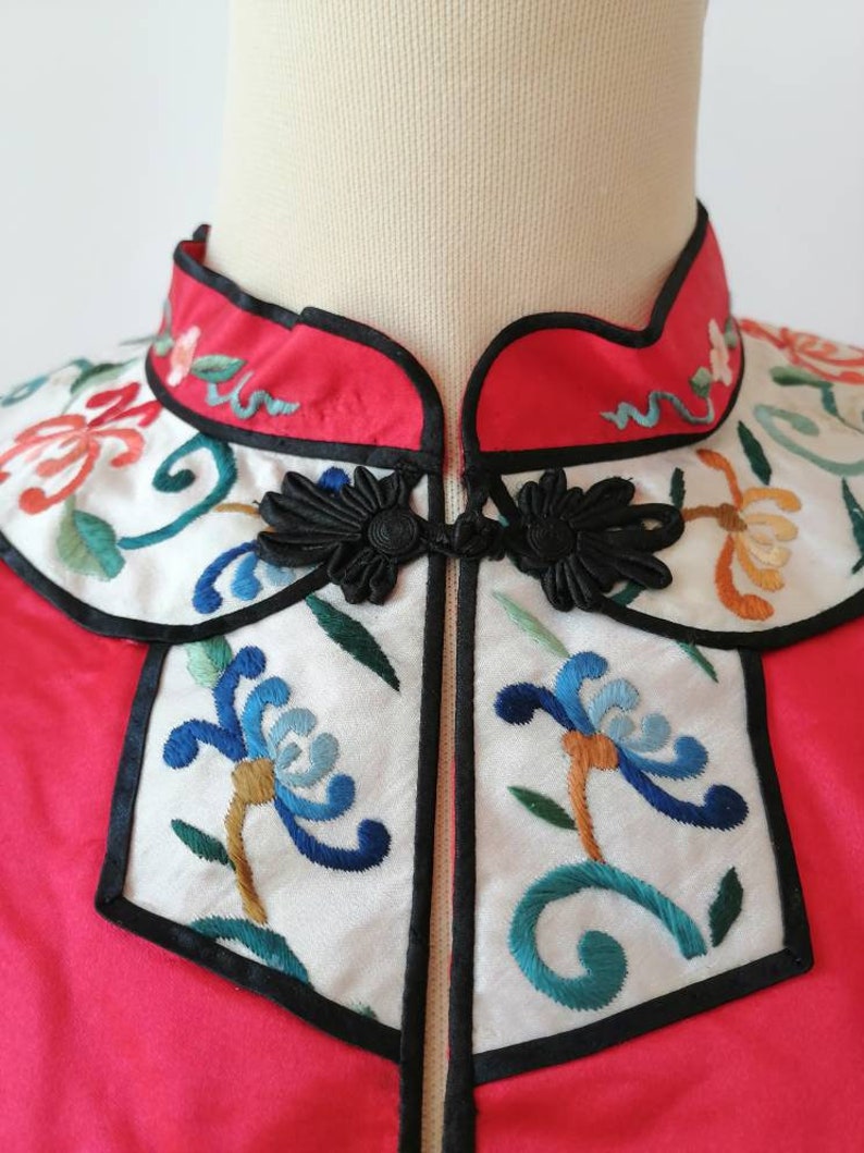 May include: A red Chinese-style jacket with a white embroidered collar and front panel. The collar and front panel feature floral designs in blue, green, orange, and white. The jacket has black frog closures.