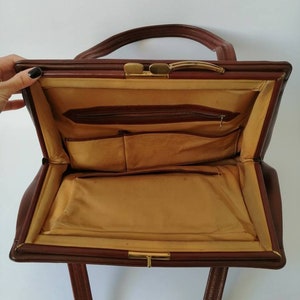 Puede incluir: Un bolso de mano de cuero marr&oacute;n con un cierre de broche dorado y un bolsillo con cremallera en el interior. El bolso tiene dos asas y un forro de tela marr&oacute;n.