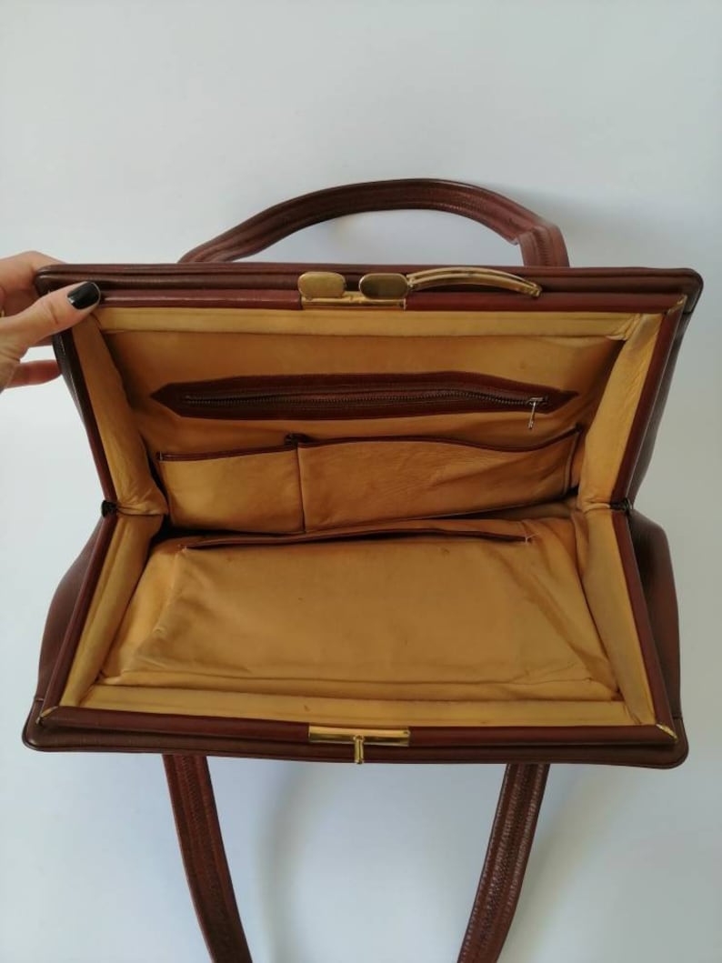 Puede incluir: Un bolso de mano de cuero marr&oacute;n con un cierre de broche dorado y un bolsillo con cremallera en el interior. El bolso tiene dos asas y un forro de tela marr&oacute;n.
