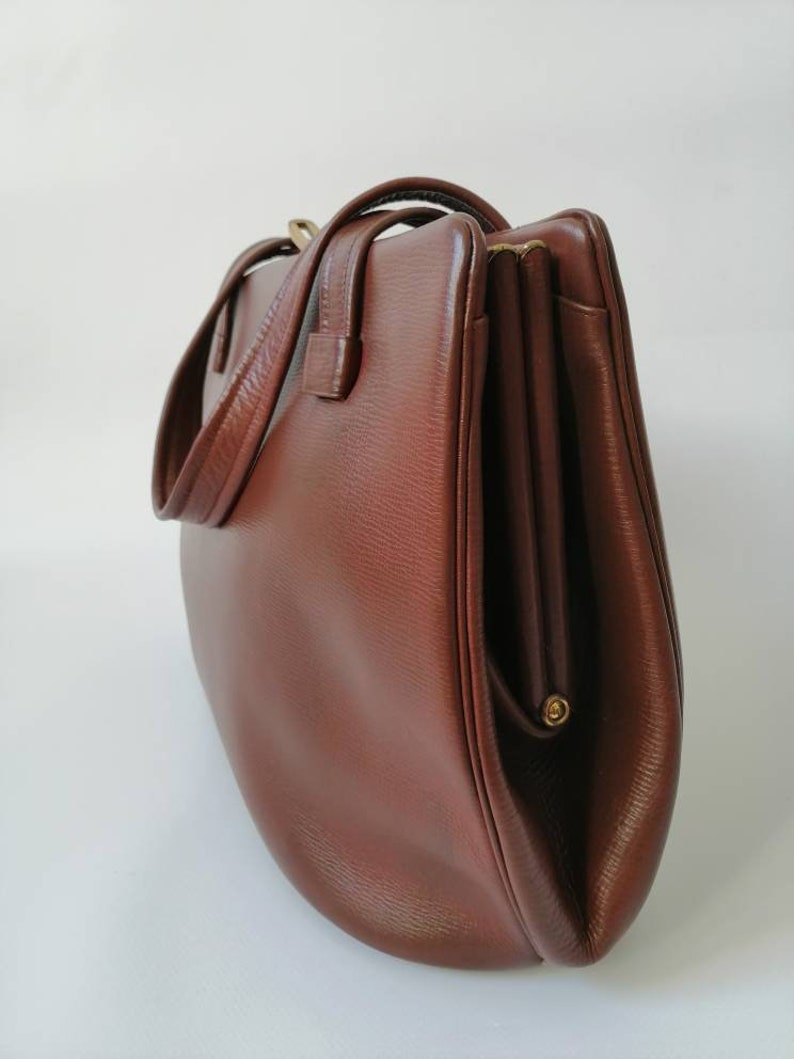 Puede incluir: Un bolso de mano de cuero marr&oacute;n con cierre de solapa y dos asas. El bolso tiene una forma ligeramente redondeada y un cierre dorado.