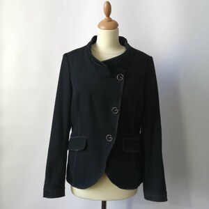 Puede incluir: Una chaqueta negra con cuello alto y un solo botón de cierre. La chaqueta tiene un diseño ligeramente asimétrico y cuenta con dos bolsillos.