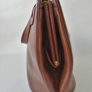 Puede incluir: Un bolso de mano de cuero marr&oacute;n con cierre de solapa y cremallera dorada. El bolso tiene una forma redondeada y un solo asa superior.