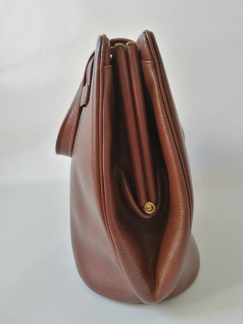 Puede incluir: Un bolso de mano de cuero marr&oacute;n con cierre de solapa y cremallera dorada. El bolso tiene una forma redondeada y un solo asa superior.