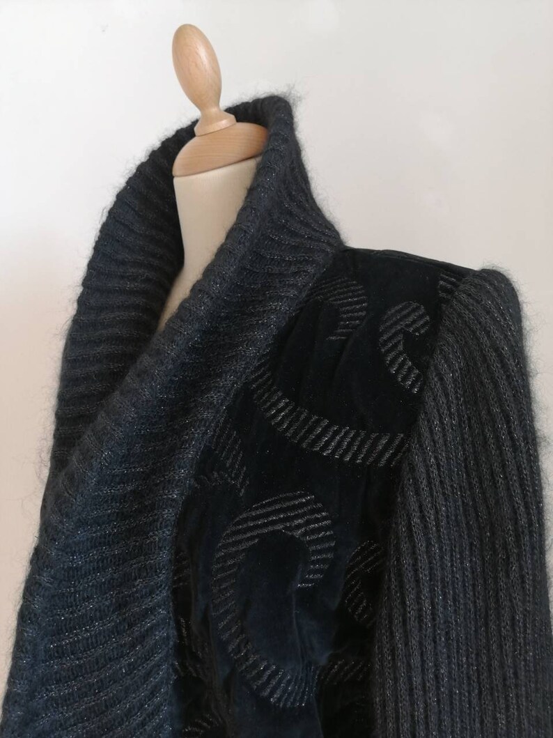 May include: A dark gray knit cardigan with a cowl neck and a black velvet panel on the right shoulder. The velvet panel has a silver embroidered design.