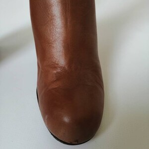 May include: Close-up of the toe of a brown leather boot. The boot has a rounded toe and a smooth leather finish.