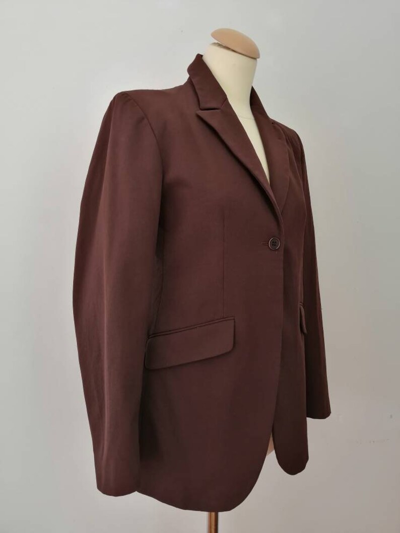 Pu&ograve; includere: Un blazer marrone con una chiusura a bottone singolo e una tasca sul lato sinistro.