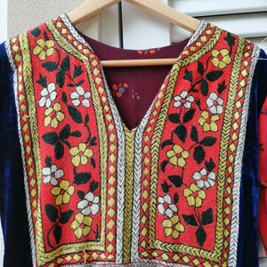 Puede incluir: Un vestido vintage rojo con ribete de terciopelo azul y bordado floral intrincado en amarillo, blanco y negro. El vestido tiene un escote en V y una falda fruncida. La etiqueta "Fru Fru Vintage" es visible en la percha.