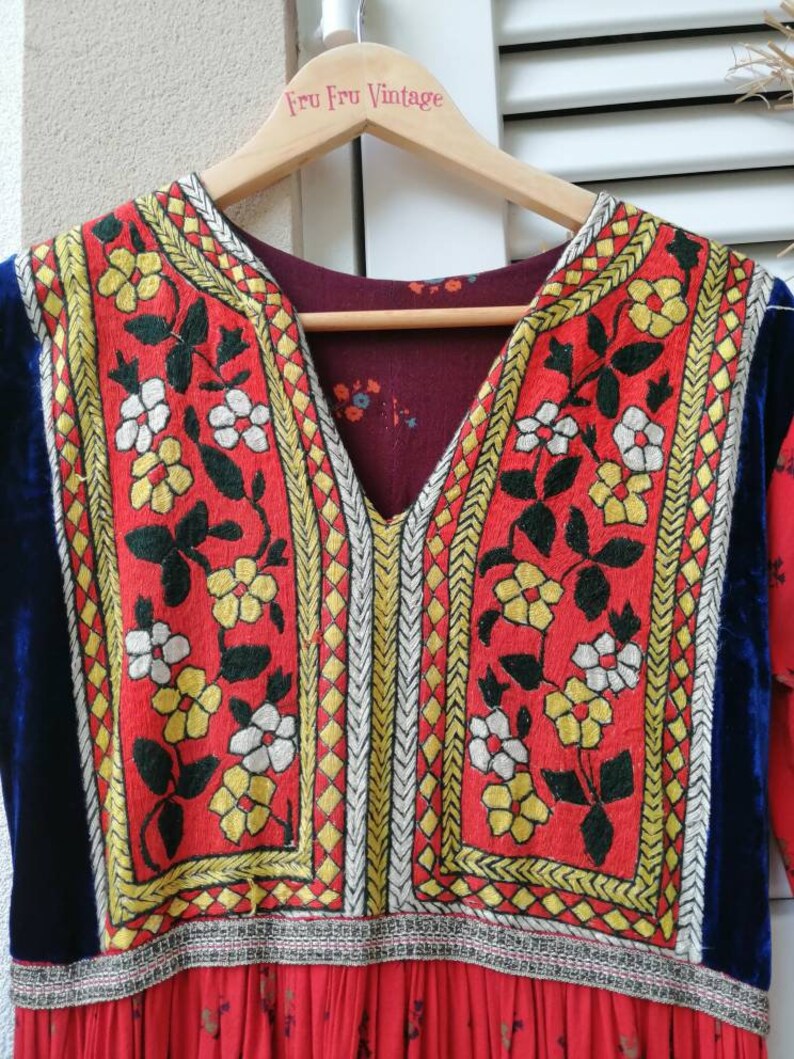 Puede incluir: Un vestido vintage rojo con ribete de terciopelo azul y bordado floral intrincado en amarillo, blanco y negro. El vestido tiene un escote en V y una falda fruncida. La etiqueta "Fru Fru Vintage" es visible en la percha.