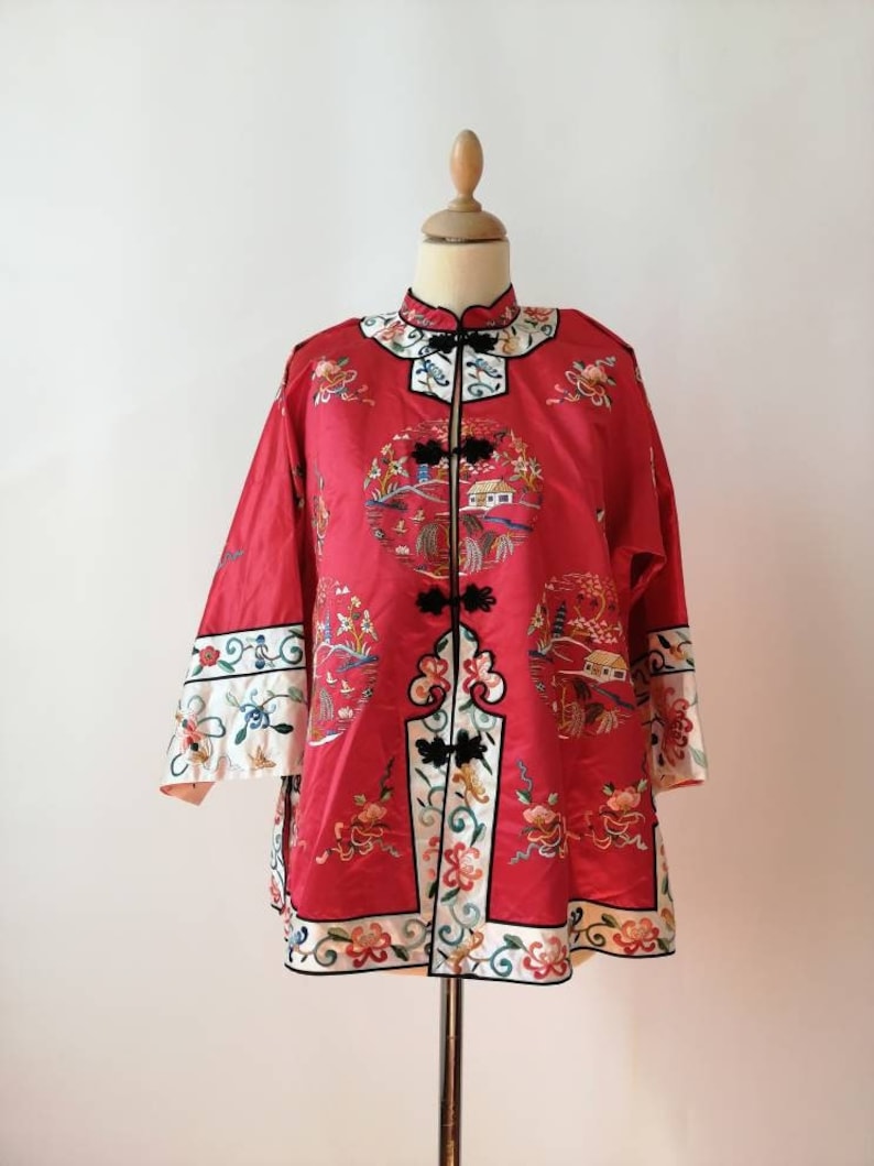 May include: A red silk jacket with intricate embroidery featuring floral patterns and a traditional Chinese landscape scene. The jacket has a mandarin collar and a front closure with black buttons.
