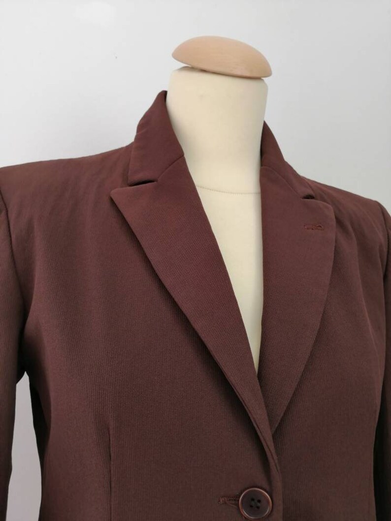 Pu&ograve; includere: Un blazer marrone con una chiusura a un solo bottone. Il blazer ha un colletto a punta e maniche lunghe.