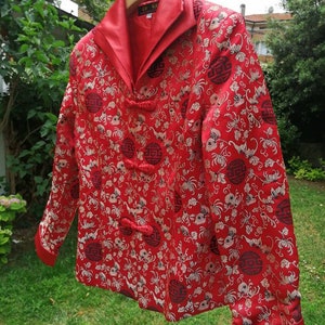 Pu&ograve; includere: Giacca rossa in broccato con colletto alla coreana e bottoni dorati. La giacca presenta un motivo floreale con un design ripetitivo di un cerchio contenente caratteri cinesi. La giacca &egrave; appesa a un appendiabiti in legno con la scritta "Free For Vintage".