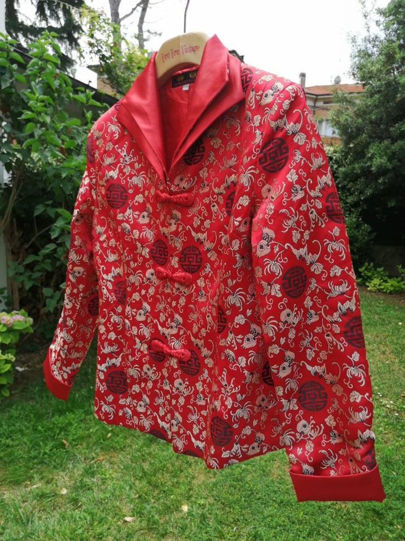 Pu&ograve; includere: Giacca rossa in broccato con colletto alla coreana e bottoni dorati. La giacca presenta un motivo floreale con un design ripetitivo di un cerchio contenente caratteri cinesi. La giacca &egrave; appesa a un appendiabiti in legno con la scritta "Free For Vintage".